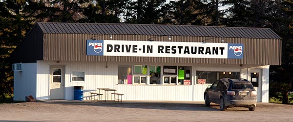 Glenboro Drive-In - a white metal-clad building with a black metal roof that overhangs the front area. A typical drive-in with order and pick-up windows. A black car sits in the parking lot. A picnic table sits to the left of the ordering area. Pepsi logos bookend a sign on the front of the building that reads "Drive-In Restaurant".