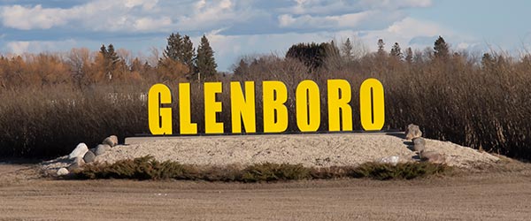 Glenboro highway sign consisting of the word Glenboro rendered in yellow, all capital letters. The letters sit atop a slight mound of gravel. a row of shrubs is in the background.